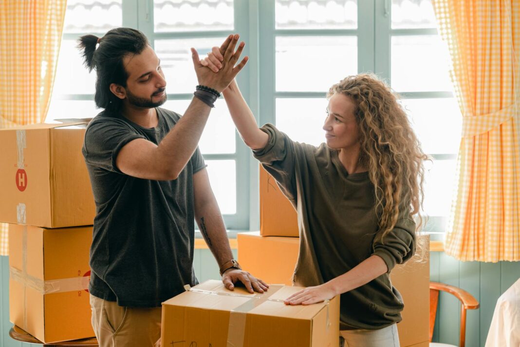 Photo by Ketut Subiyanto Happy woman in casual wear standing near heap of cardboard boxes and giving high five to ethnic boyfriend with ponytail showing agreement while looking at each other
