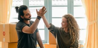 Happy woman in casual wear standing near heap of cardboard boxes and giving high five to ethnic boyfriend with ponytail showing agreement while looking at each other