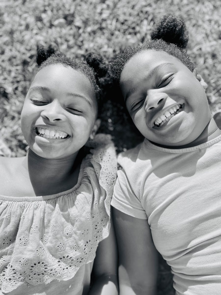 Two happy children with natural hair laying on grass and smiling in a black and white portrait.