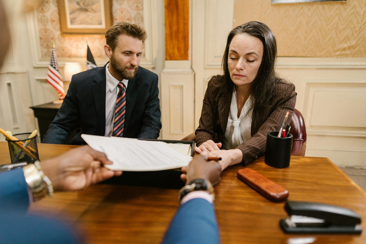 Three professionals exchange legal documents during a formal meeting at a conference table, representing client acquisition strategies for law firms.