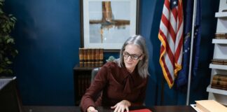 Professional attorney with glasses writes in red folder at wooden desk in law office with American flag and legal books, illustrating legal directory professional profiles.