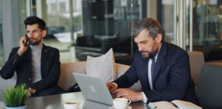 Two businessmen work in a modern glass-walled office, one at a laptop with documents, representing law firm professionals using digital strategies for client acquisition.