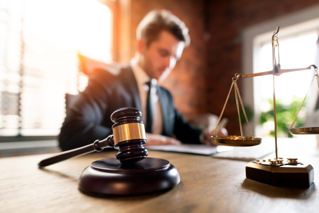 Traditional symbols of justice frame modern legal practice and directory technology in professional law office Wooden gavel and brass scales of justice on a desk with a lawyer working at a computer in the background, representing modern legal practice and professional directories.