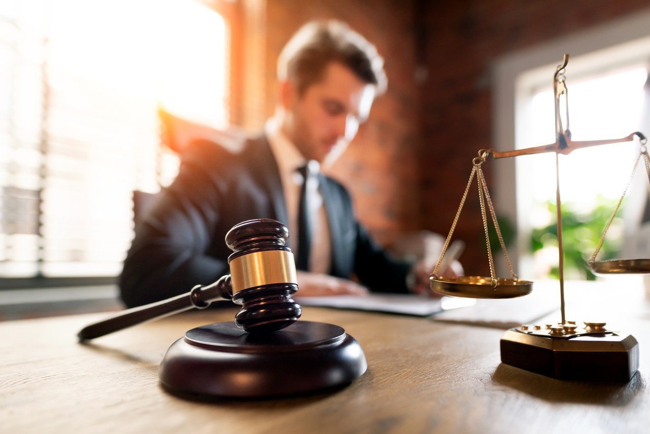 Wooden gavel and brass scales of justice on a desk with a lawyer working at a computer in the background, representing modern legal practice and professional directories.