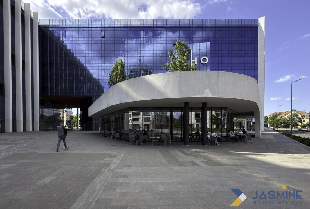 Modern commercial architecture exemplifies the physical infrastructure supporting business directories and professional connectivity Contemporary glass office building with curved white canopy and outdoor cafe plaza in Timișoara, illustrating the commercial infrastructure where businesses maintain directory listings.