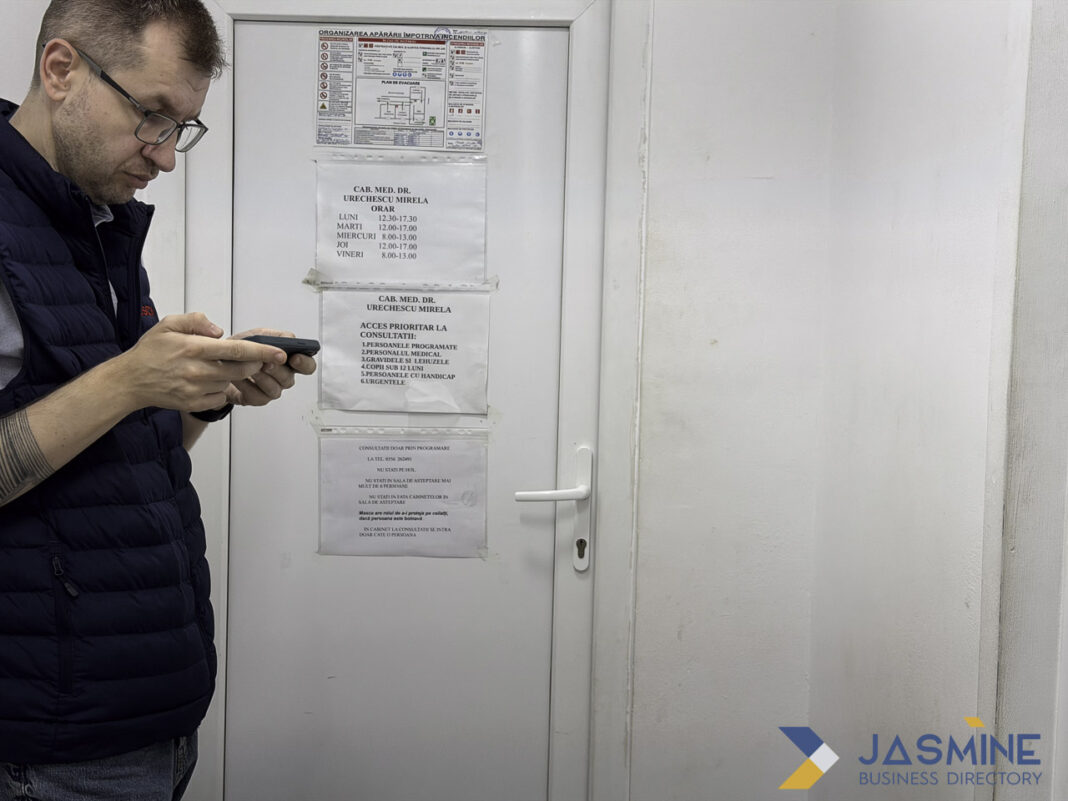 Patient accessing healthcare information on smartphone illustrates digital physician directory usage Man wearing glasses uses smartphone in medical office waiting area with appointment schedules visible behind him, representing digital access to physician directories and healthcare information.