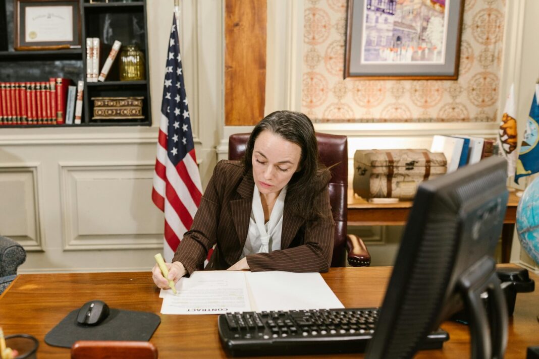 Attorney workspace representing legal professionals listing their practice in state law firm directory Professional woman in pinstripe suit working at traditional office desk with American flag, representing attorneys listing their firms in legal directories.