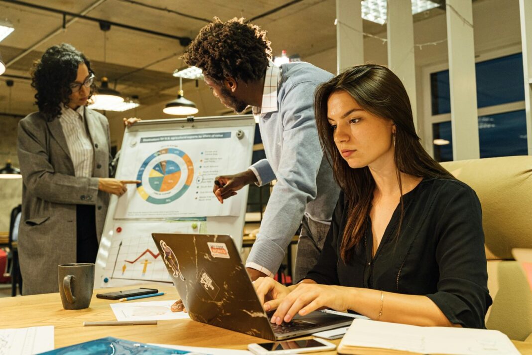 Professional woman develops business description at laptop in collaborative workspace Woman types on laptop at wooden desk in modern office while colleagues discuss charts on whiteboard, illustrating the process of writing business descriptions.