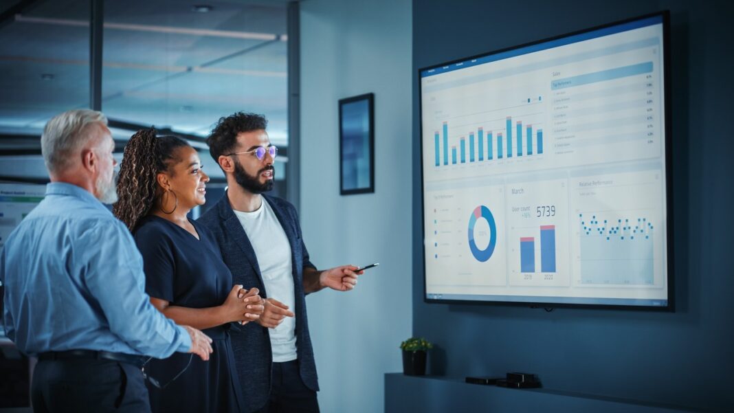 Business team analyzes performance metrics illustrating shift from lead generation to demand capture strategies Three business professionals review data visualizations on a large office screen, analyzing performance metrics central to modern B2B demand capture strategies.
