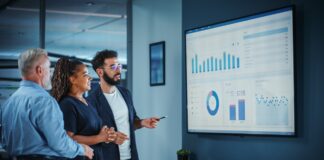 B2B Marketing Trends: From Lead Gen to Demand Capture Three business professionals review data visualizations on a large office screen, analyzing performance metrics central to modern B2B demand capture strategies.