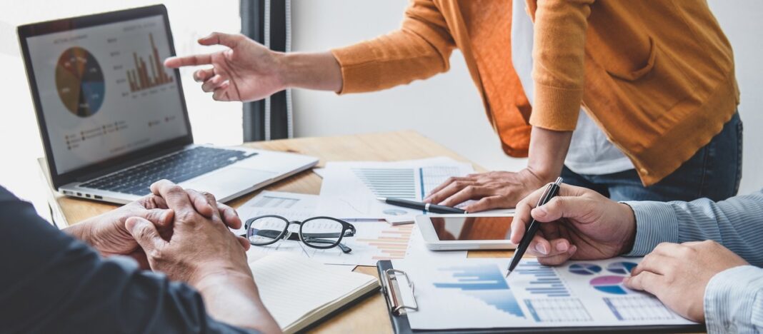 Business professionals review data analytics to illustrate citation profile audit strategies Three business professionals collaborate around a table with laptop, charts, and documents, representing the data review process essential for auditing citation profiles.