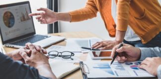 How to Audit Your Citation Profile for AI Visibility Three business professionals collaborate around a table with laptop, charts, and documents, representing the data review process essential for auditing citation profiles.
