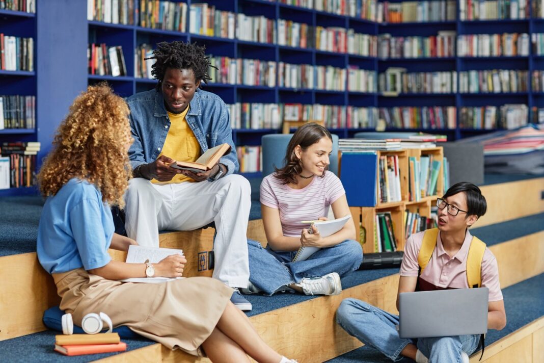 Library study group exemplifies effective face-to-face communication and collaborative dialogue Four students sit in a circle on a wooden library platform engaged in discussion, illustrating effective interpersonal communication and collaborative dialogue.
