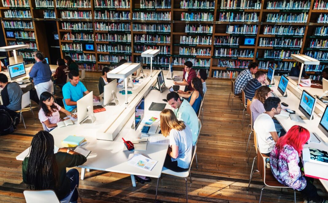 Library study environment illustrates research and information discovery processes central to effective keyword strategy. Students work at white desks with laptops in a library surrounded by bookshelves, representing the research process involved in finding effective keywords.