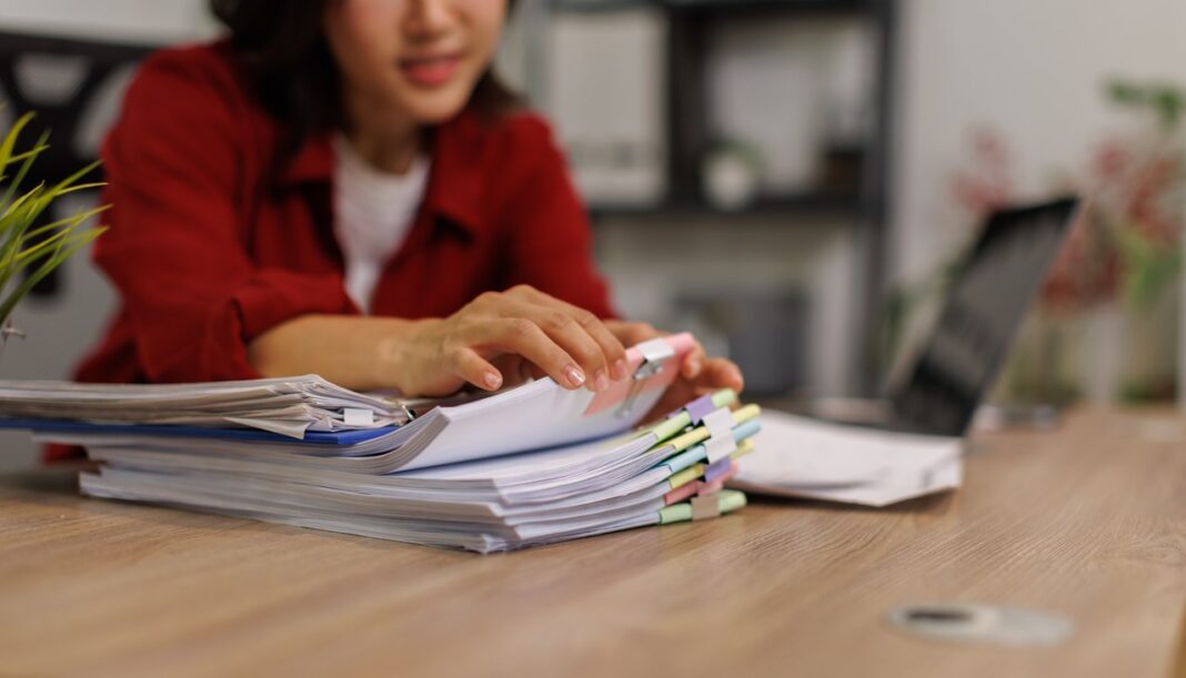 Office worker in red blazer organizing stacked paper documents with colored tabs at wooden desk, representing systematic categorization methods used in web directories.