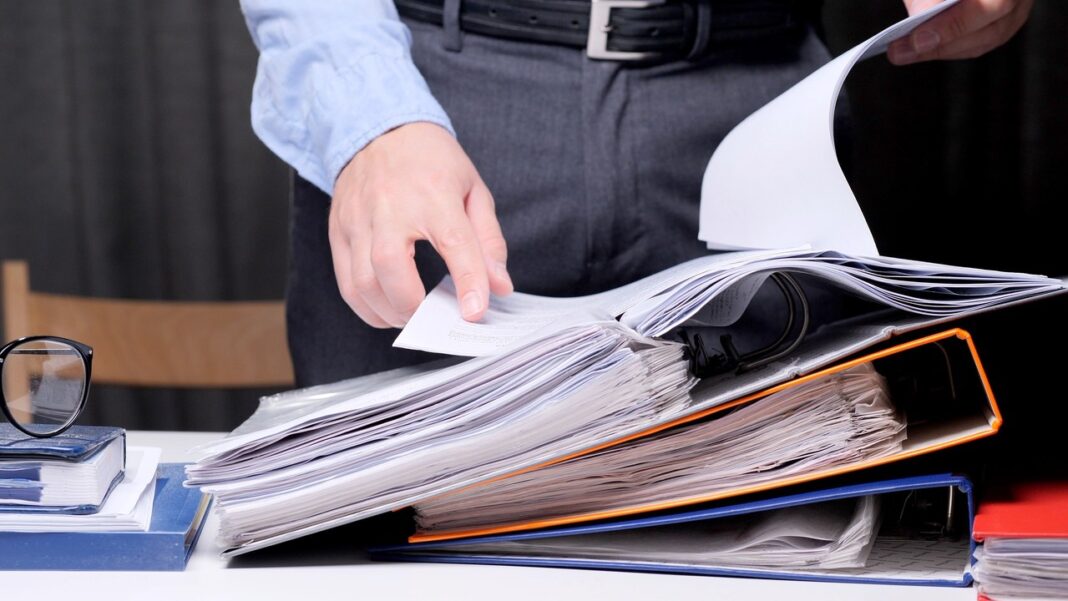Business professional organizing physical documents illustrates traditional listing management challenges Business professional reaches for documents in overstuffed ring binders at desk with eyeglasses and folders, representing manual listing management challenges.