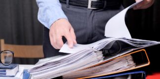 How to Manage All My Listings? Business professional reaches for documents in overstuffed ring binders at desk with eyeglasses and folders, representing manual listing management challenges.