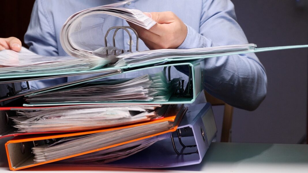 Colorful ring binders illustrate systematic organization and document management for industry directories. Person holding open a ring binder above stacked colorful overstuffed binders on a desk, representing organized directory and information management systems.