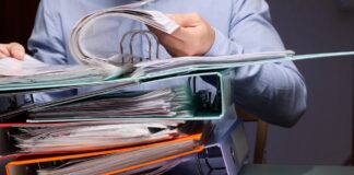 A Curated List of Industry Directories Person holding open a ring binder above stacked colorful overstuffed binders on a desk, representing organized directory and information management systems.