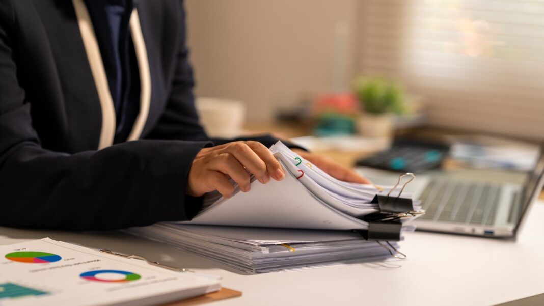 Workspace scene illustrating research and document analysis for practitioner and agency directories. Person at desk handling stacked paper documents with laptop nearby, representing the methodical research process for practitioner and agency verification.
