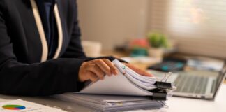 Other Practitioner & Agency Research Person at desk handling stacked paper documents with laptop nearby, representing the methodical research process for practitioner and agency verification.