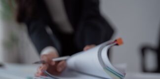 Stack of papers and magazines on an office desk with a person reviewing documents in the background, representing content organization and duplicate material management.