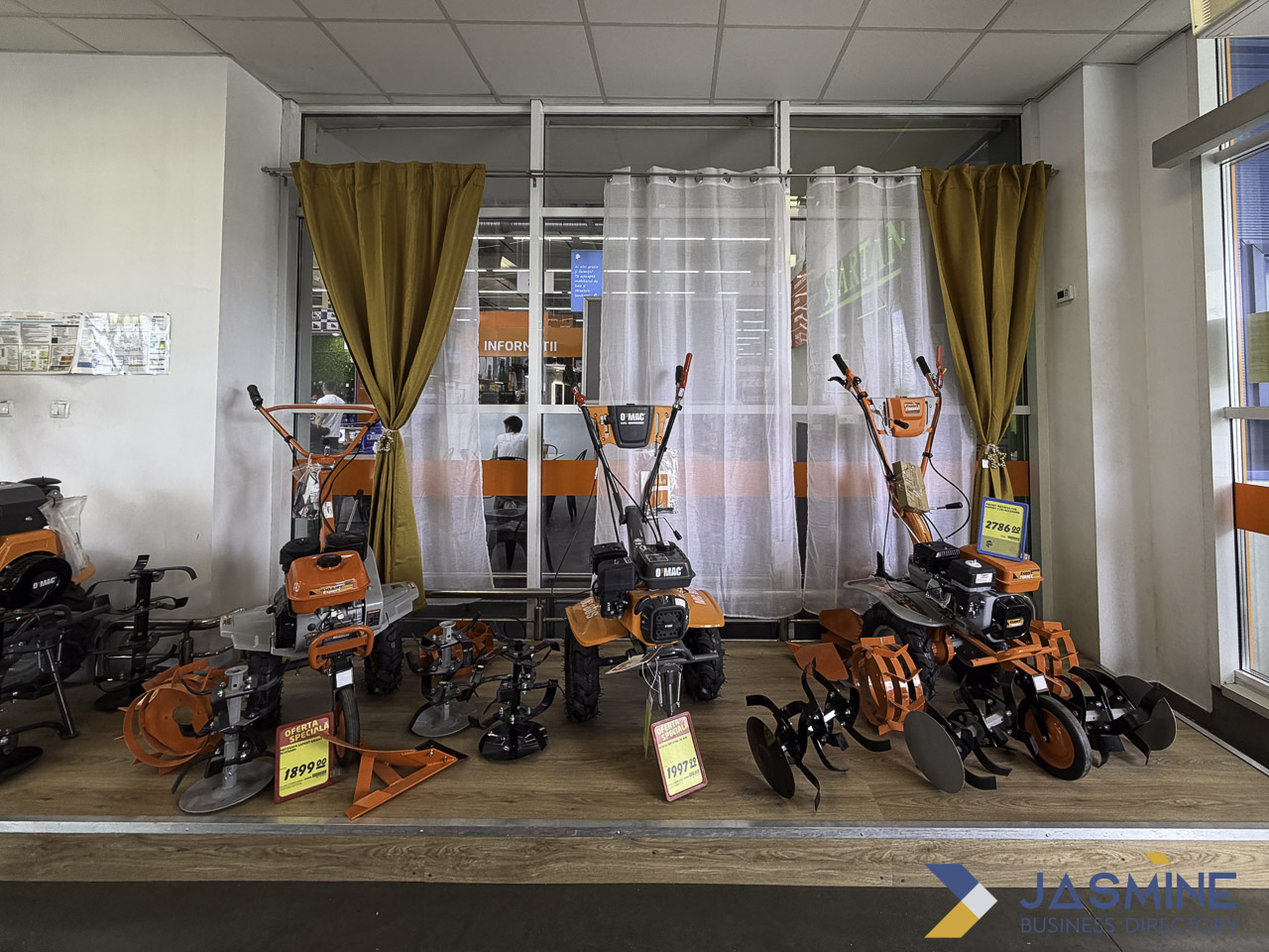 Orange and black agricultural tillers displayed in a showroom with glass partitions and curtains, illustrating retail product presentation for local business discovery.