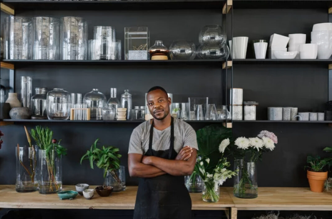 Artisan entrepreneur embodies slow marketing's focus on craftsmanship and deliberate growth Artisan business owner in dark apron stands at wooden counter in organized workshop with shelving and flowers, representing the deliberate craftsmanship central to slow marketing principles.