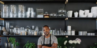 The “Slow Marketing” Movement: Building Long-Term Value Over Quick Wins Artisan business owner in dark apron stands at wooden counter in organized workshop with shelving and flowers, representing the deliberate craftsmanship central to slow marketing principles.