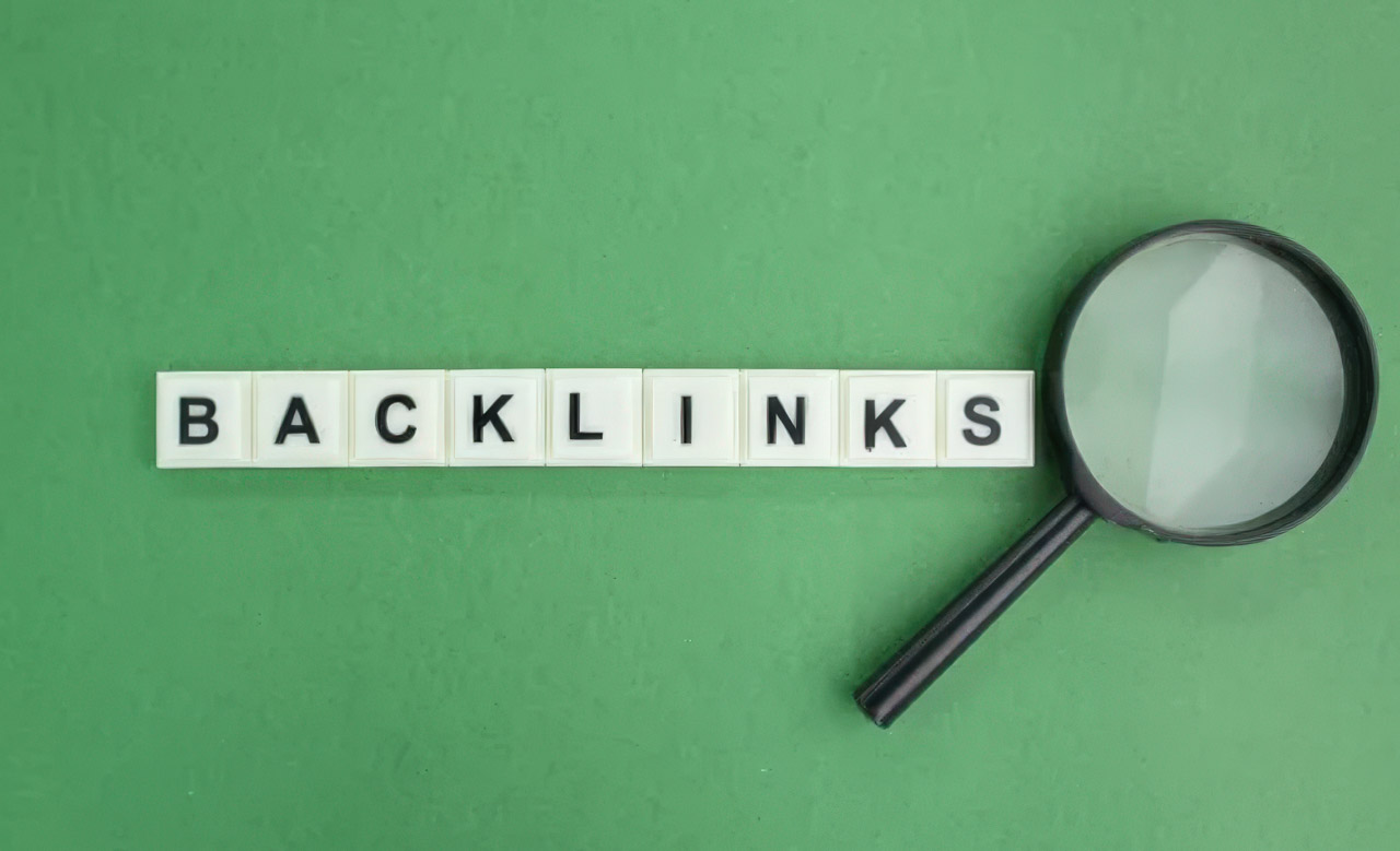 White letter tiles spelling BACKLINKS on green surface beside a magnifying glass, representing search engine optimization and link building strategies.