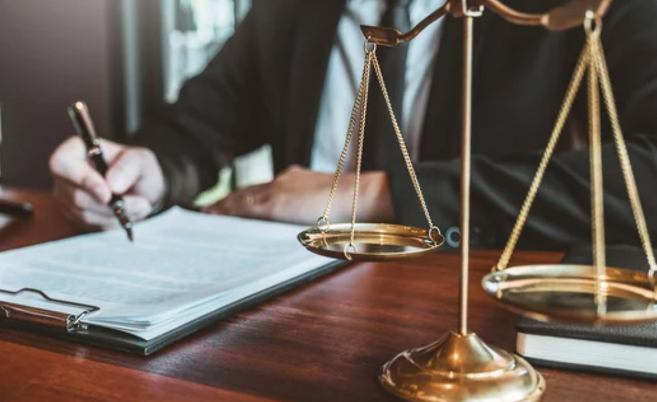 Brass balance scale of justice on a wooden desk with a lawyer writing documents in the background, illustrating professional legal directory profiles.