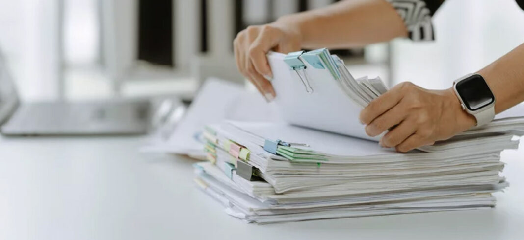 Organizing paperwork and documentation for business directory submissions Hands holding white papers clipped with green and pink binder clips above document stacks on a white desk, representing the documentation needed for business directory listings.