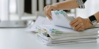 Hands holding white papers clipped with green and pink binder clips above document stacks on a white desk, representing the documentation needed for business directory listings.