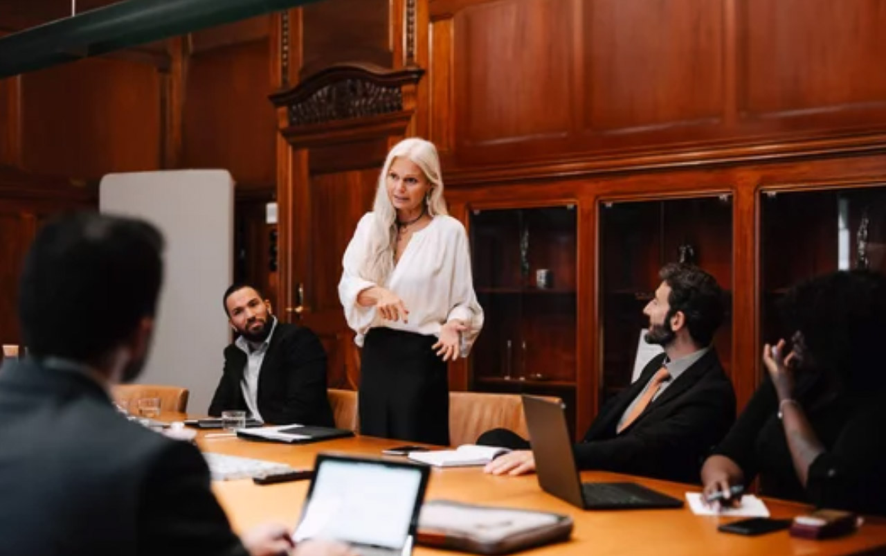 A businesswoman stands presenting to seated colleagues around a conference table with laptops, illustrating the collaborative research process for selecting business directories.