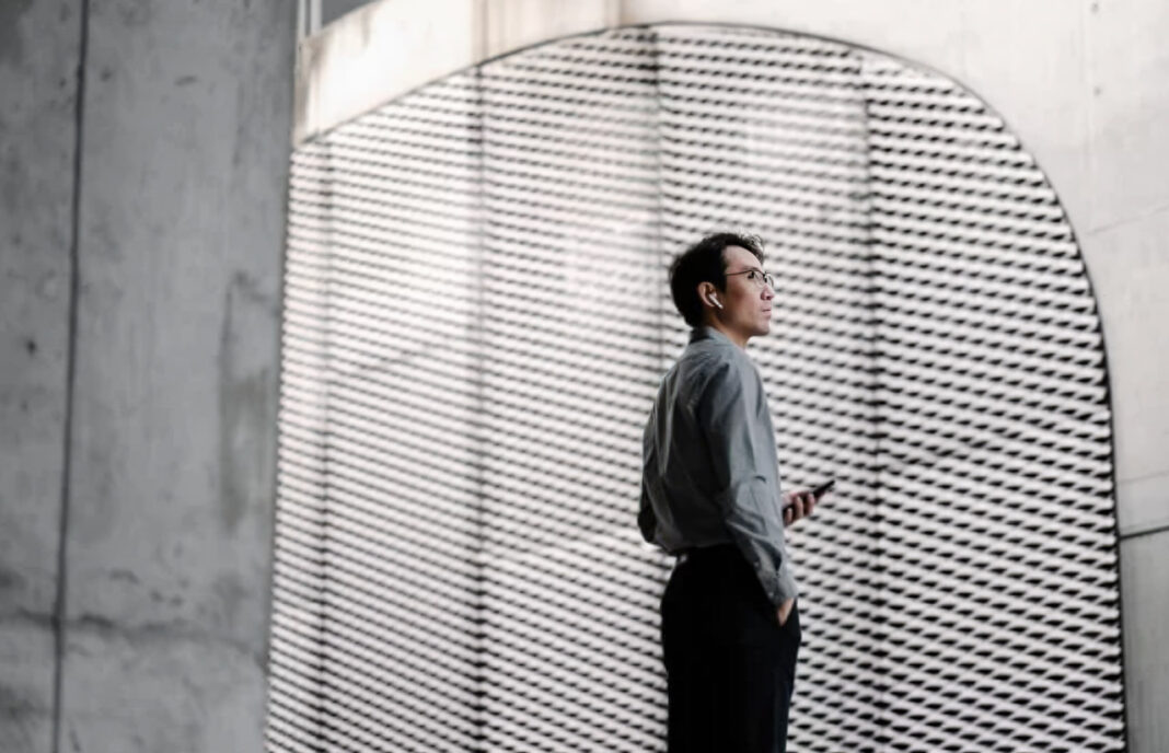 Modern business professional in architectural setting represents entity-first business identity and establishment A business professional in grey shirt stands in profile within a modern minimalist space with perforated wall panels, representing contemporary business identity and establishment.