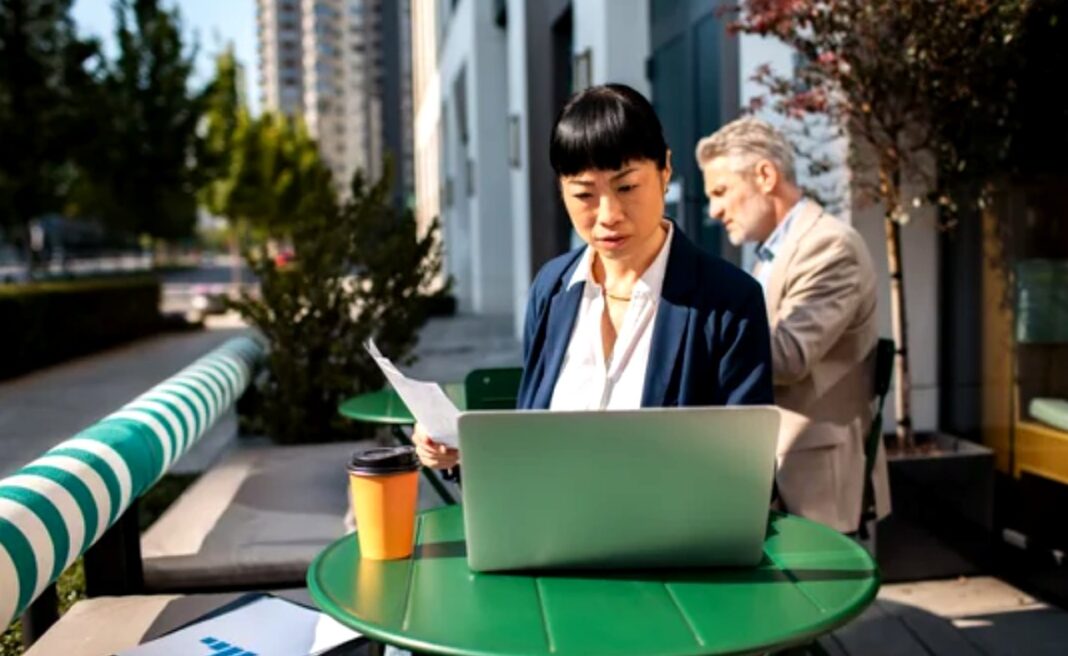 Modern professionals embrace flexible outdoor workspaces to enhance business visibility and productivity in 2025 Woman in business attire working on laptop at outdoor table with urban skyline backdrop, illustrating modern professional visibility and digital business practices.