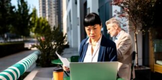 Get Your Business Found in 2027 Woman in business attire working on laptop at outdoor table with urban skyline backdrop, illustrating modern professional visibility and digital business practices.