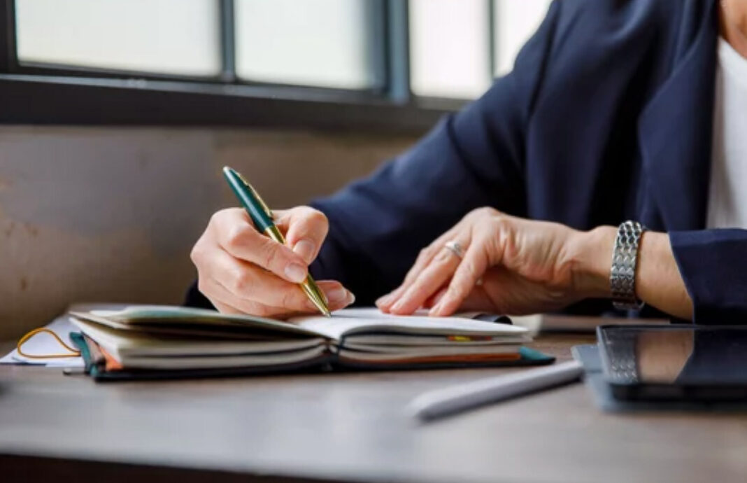 Planning and documentation essential for writing content that serves both human readers and AI systems Close-up of hands in business attire writing with a gold pen in a notebook at a wooden desk, illustrating the planning process for creating dual-audience content.