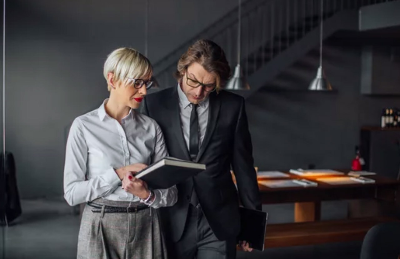 Two business professionals in suits review an open document together at an office desk, illustrating how small business owners research and consult directory resources.