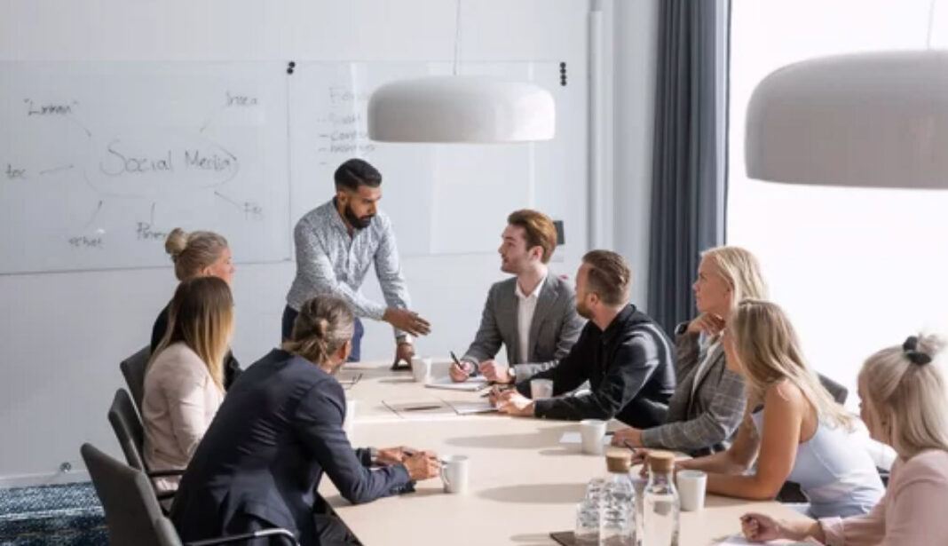 Corporate team collaborates on social media strategy, illustrating marketing's shift toward networked engagement. A presenter stands before seated colleagues around a conference table in a modern office, with a whiteboard showing 'Social Media' behind them, illustrating contemporary collaborative marketing approaches.
