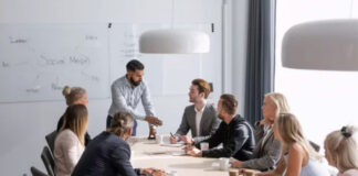 From Funnel to Mention: Marketing’s New Direction A presenter stands before seated colleagues around a conference table in a modern office, with a whiteboard showing 'Social Media' behind them, illustrating contemporary collaborative marketing approaches.
