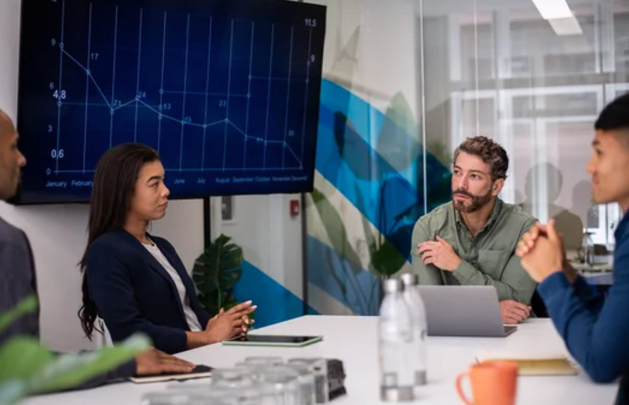 Four professionals review data on a wall screen during a conference room meeting, illustrating the strategic planning behind directory-based SEO optimization.