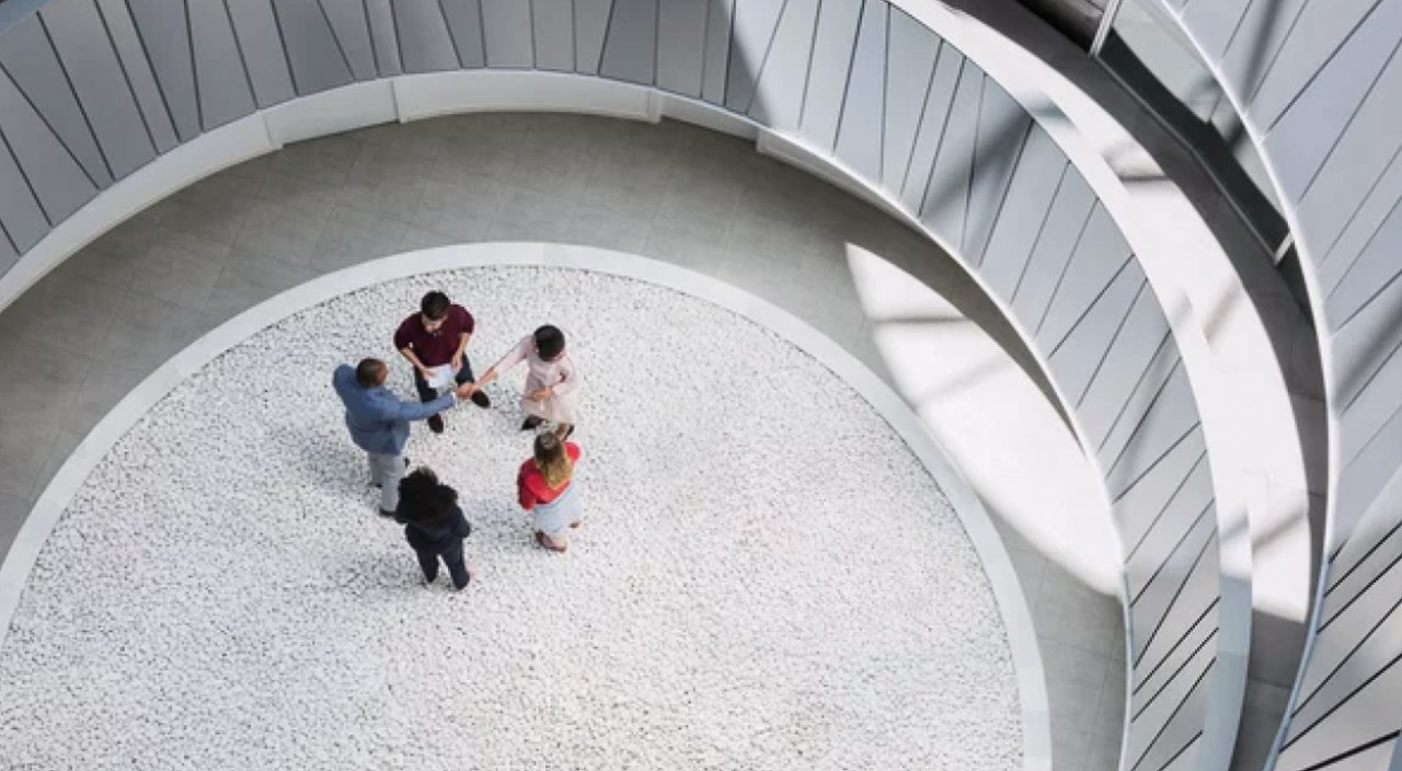 Five business professionals stand in a circle within a modern spiral atrium, symbolizing collaborative infrastructure and systematic trust-building in organizational frameworks.