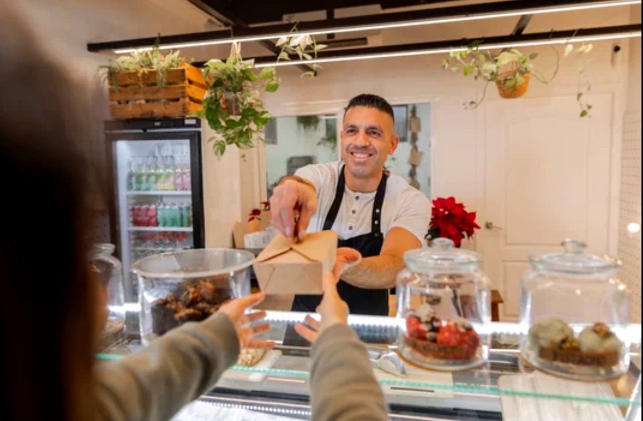 Cafe worker in black apron hands takeout box to customer at bakery counter with pastries in display case, illustrating personal service at local establishments.