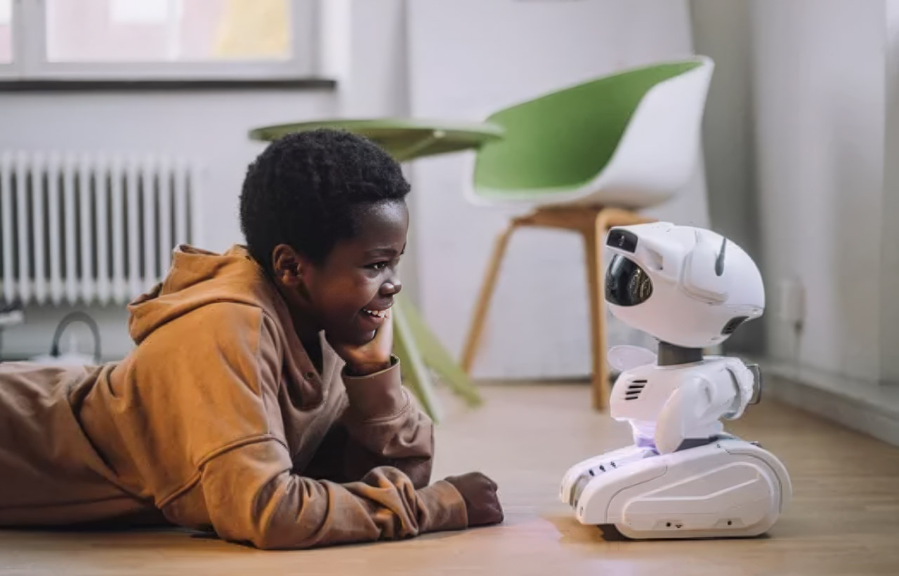 A child in an orange hoodie lies on a wooden floor interacting with a white humanoid robot, representing the future of conversational AI technology.