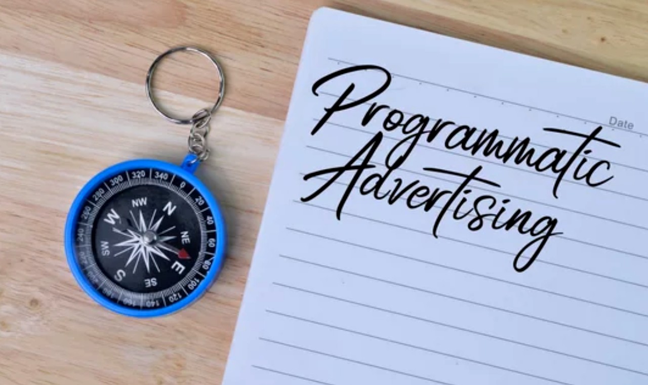 A compass keychain and notepad with 'Programmatic Advertising' handwritten text rest on a wooden surface, symbolizing strategic direction in digital advertising campaigns.