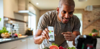Video Advertising Evolution: Short-Form, Shoppable, and Searchable Person filming smartphone screen displaying health content while standing behind fresh produce on kitchen counter, illustrating modern short-form video advertising techniques.