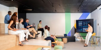 Predictive Customer Service: The New Marketing Frontline A woman presents data analytics on a large screen to seated business professionals in a modern office, illustrating predictive customer service strategies.