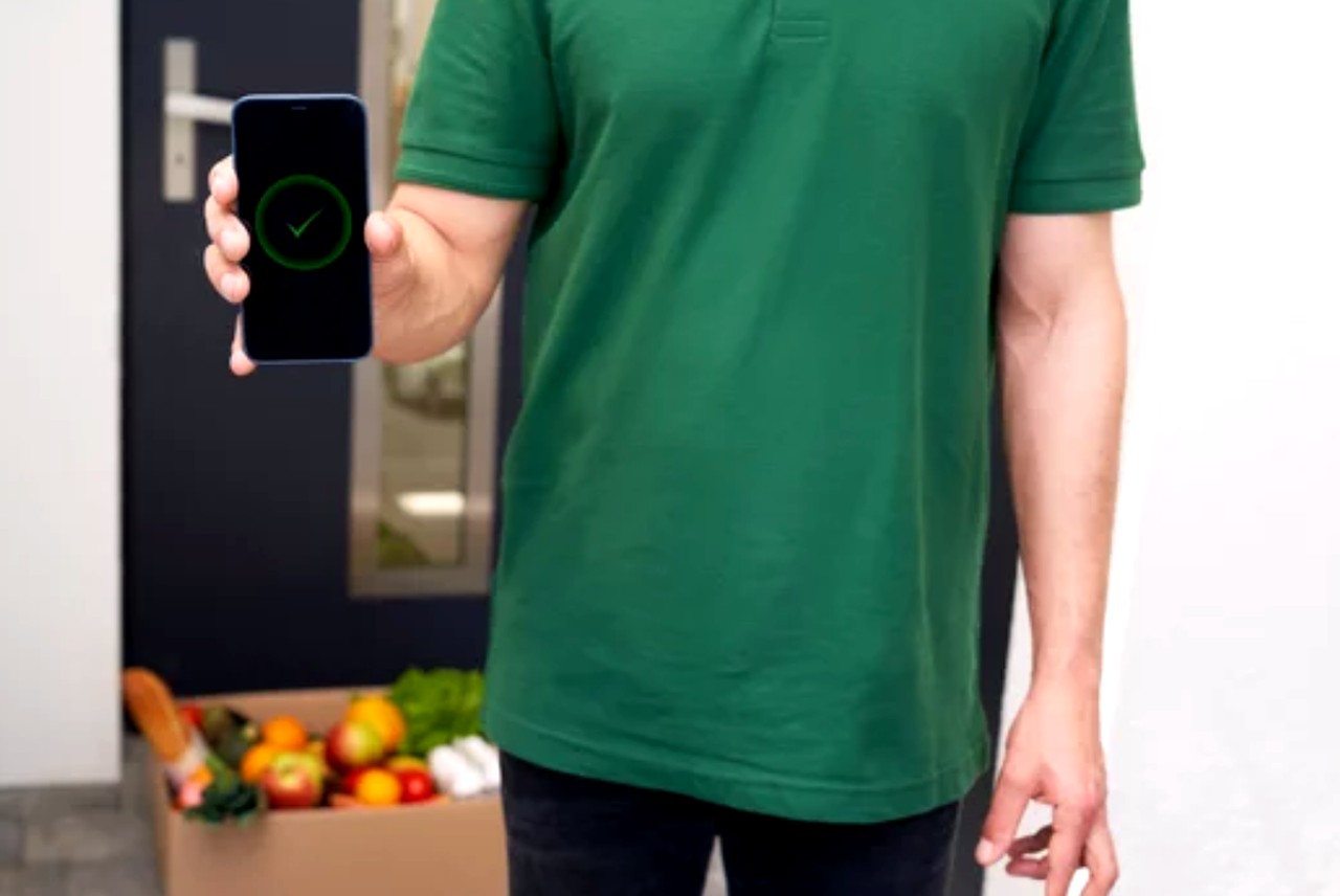 Delivery person in green polo holds smartphone showing confirmation checkmark with produce box nearby, illustrating how consistent contact information enables reliable service delivery.