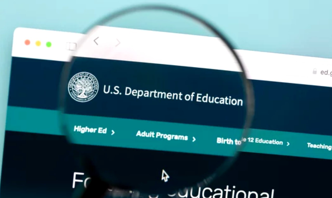 Magnifying glass focusing on U.S. Department of Education website header, representing examination of federal policies that shape student funding opportunities.